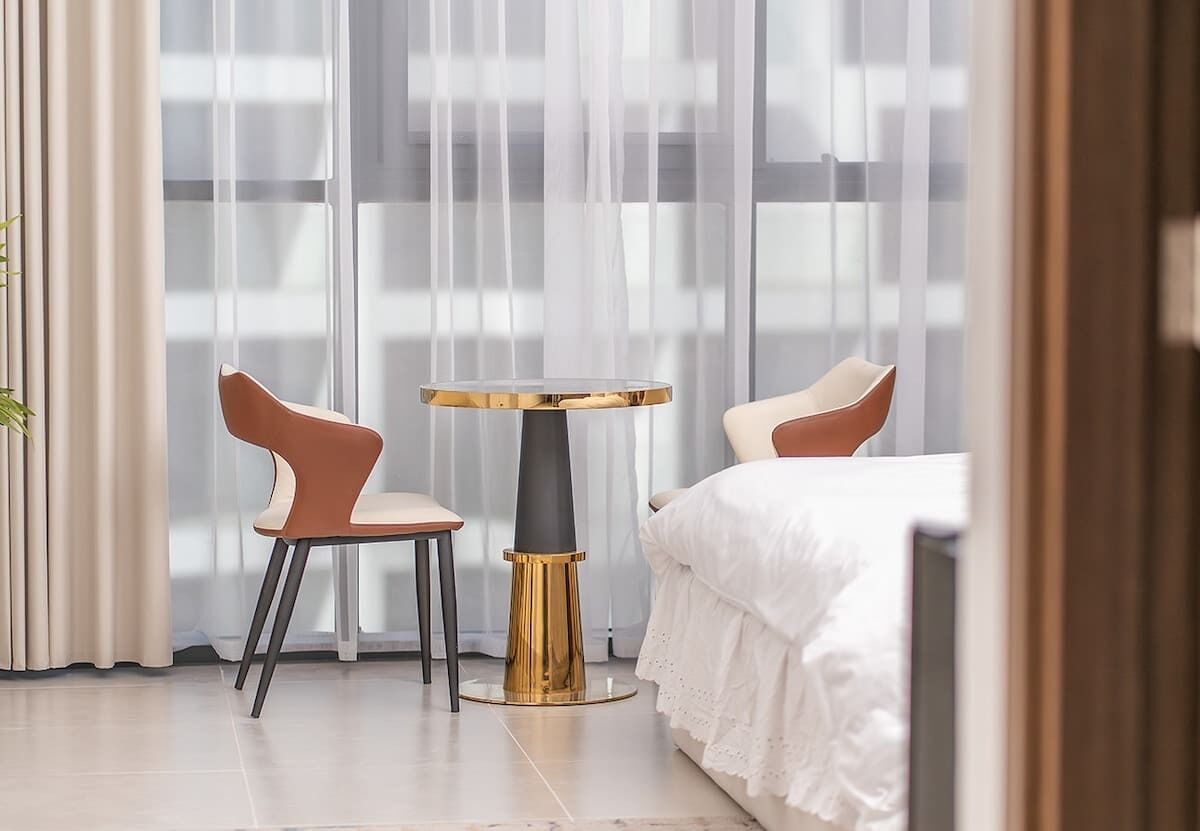A hotel room with a bed, two chairs, a small round table, and sheer curtains.