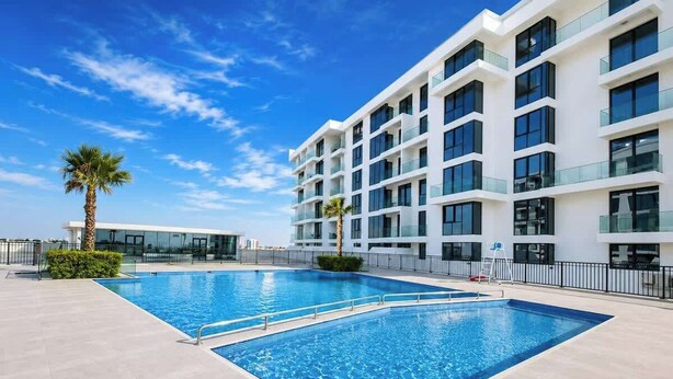 Un hotel moderno con piscina, varios balcones y un cielo azul despejado.