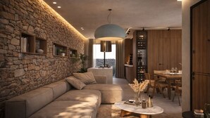 A modern living room with a stone wall, a sectional sofa, a round coffee table, and a dining area with wooden cabinets and a chandelier. - Family Villa with Pool, Fire Pit, Sports Courts - Perfect for Groups (Pigi)