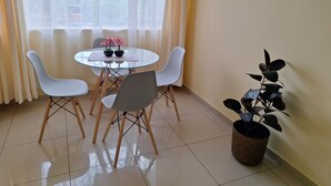Un área de comedor con una mesa redonda de vidrio, cuatro sillas blancas, dos plantas en macetas y una ventana con cortinas de gasa.