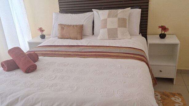 Una cama bien hecha con una colcha estampada, dos almohadas y un cojín decorativo. Hay dos mesitas de noche con pequeños jarrones de flores.
