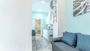 A modern living room with a grey sofa, a small rug, and a wooden floor. A kitchen with white cabinets and a wooden countertop is visible in the background. - Shopping Paradise 9BR For 22 People (Vienna)