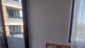 A single bed with a wooden frame and a grey mattress in a room with a large window and a view of a building outside.