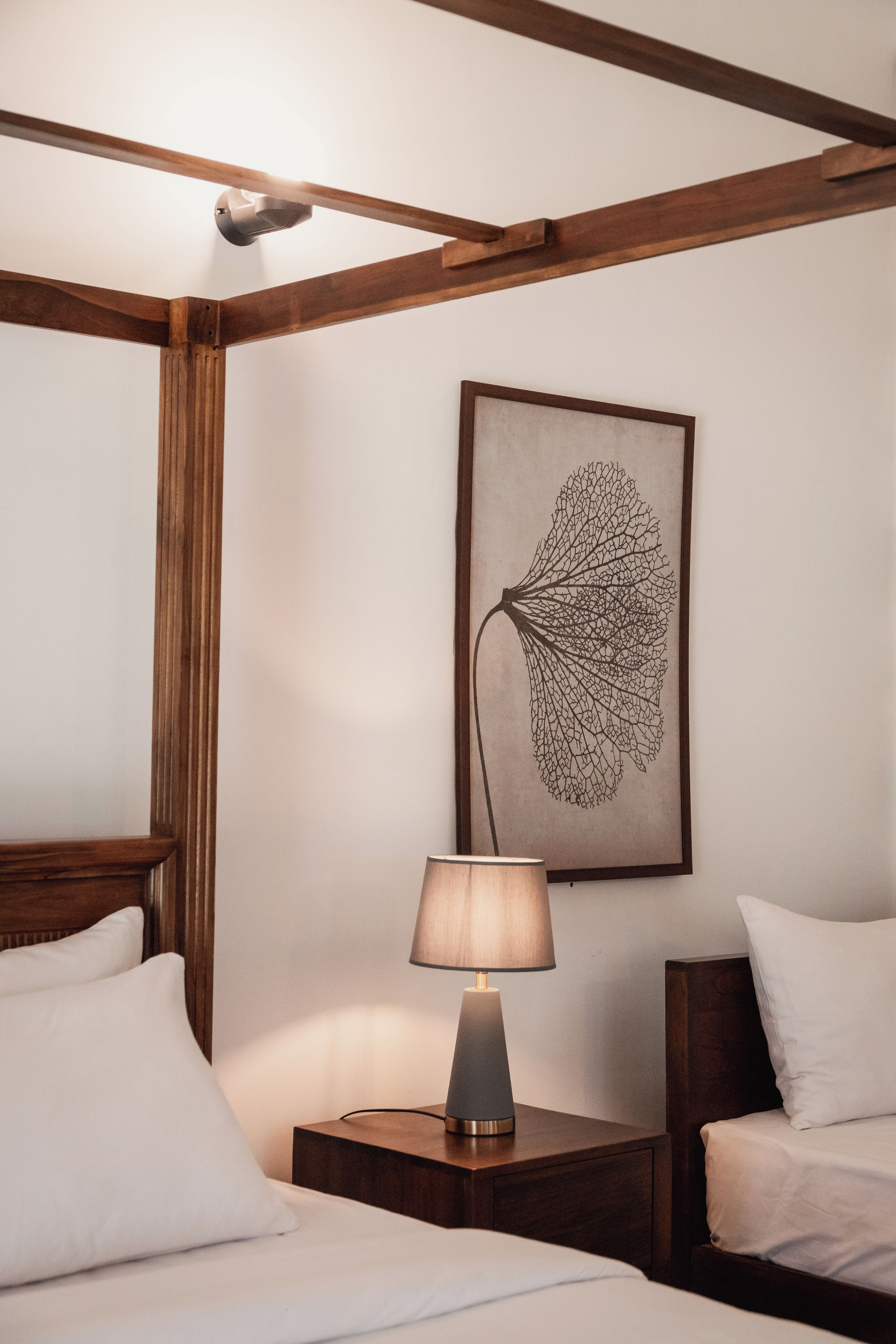 A hotel room with a wooden ceiling, a framed leaf artwork on the wall, a bedside lamp, and two beds with white bedding.