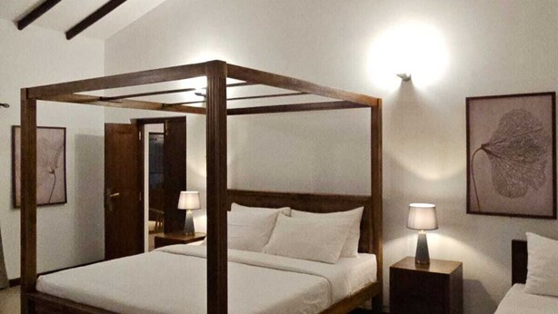 Un lit à baldaquin avec une structure en bois et une literie blanche, une table de chevet avec une lampe, et une œuvre d’art encadrée accrochée au mur.