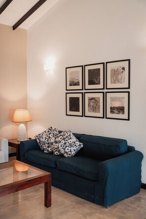 Een woonkamer met een donkerblauwe bank, een glazen salontafel, een lamp en zes ingelijste foto's aan de muur.