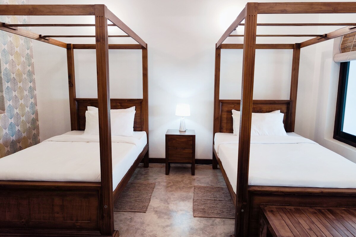Two four-poster beds with wooden frames and white linens, a small bedside table with a lamp, and a window with a view of the outside.