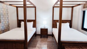 Dos camas con dosel y estructura de madera, cubiertas con sábanas blancas, una mesita de noche con lámpara y una ventana que ofrece vistas al exterior.