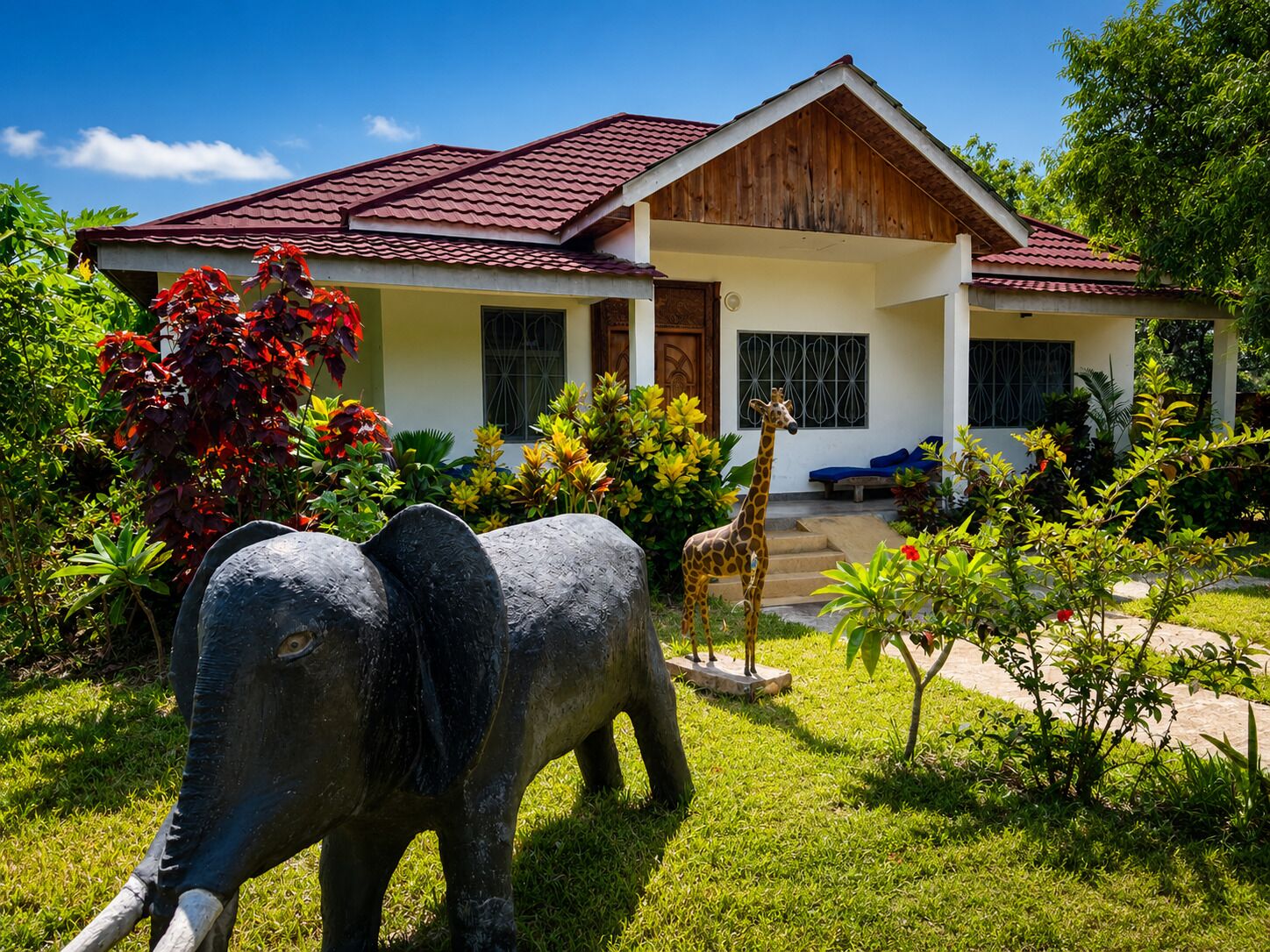 A house with a red-tiled roof, a front porch, and a garden with a giraffe and an elephant statue.