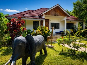 A house with a red-tiled roof, a front porch, and a garden with a giraffe and an elephant statue. - Diamond Coast Villa and Apartments (Paje)