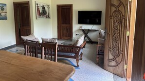 A living room with a wooden dining table, two wooden chairs, a sofa, a TV mounted on the wall, and a decorative wooden door. - Diamond Coast Villa and Apartments (Paje)