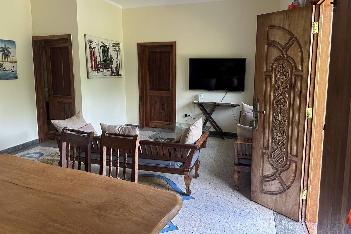 A living room with a wooden dining table, two wooden chairs, a sofa, a TV mounted on the wall, and a decorative wooden door.