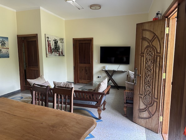 Una sala de estar con una mesa de comedor de madera, dos sillas de madera, un sofá, un televisor montado en la pared y una puerta decorativa de madera.