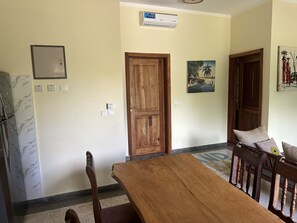 A dining area with a wooden table and chairs, a wooden door, a wall-mounted air conditioner, and a small picture on the wall. - Diamond Coast Villa and Apartments (Paje)