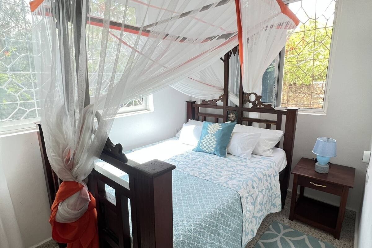 A bedroom with a four-poster bed, a bedside table with a lamp, and a window with a view of greenery.