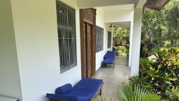 A covered outdoor seating area with two blue lounge chairs, a wooden bench, and a small table. - Diamond Coast Villa and Apartments (Paje)