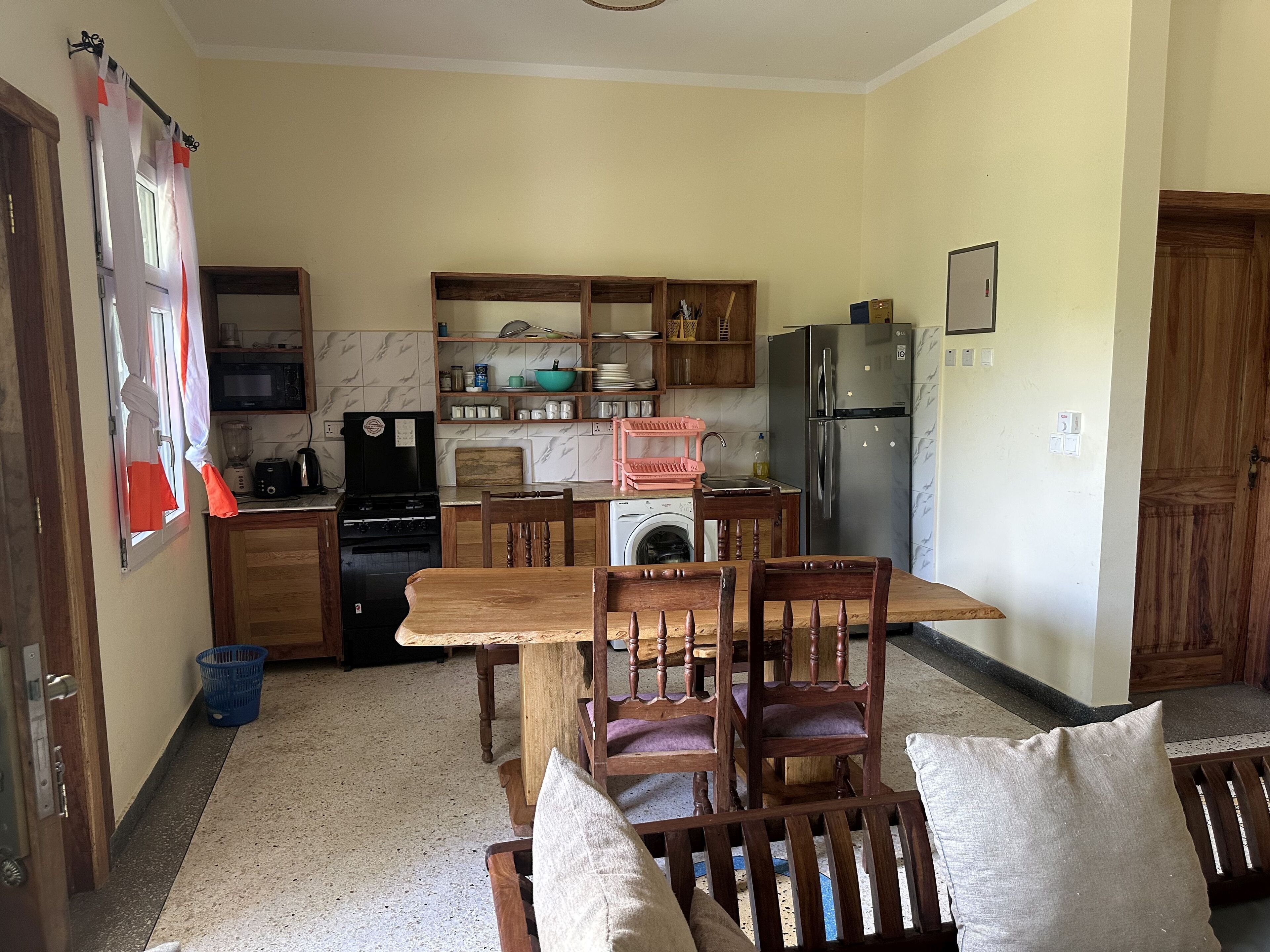 A compact kitchen with wooden cabinets, a washing machine, a refrigerator, and a dining area with wooden chairs and a table.