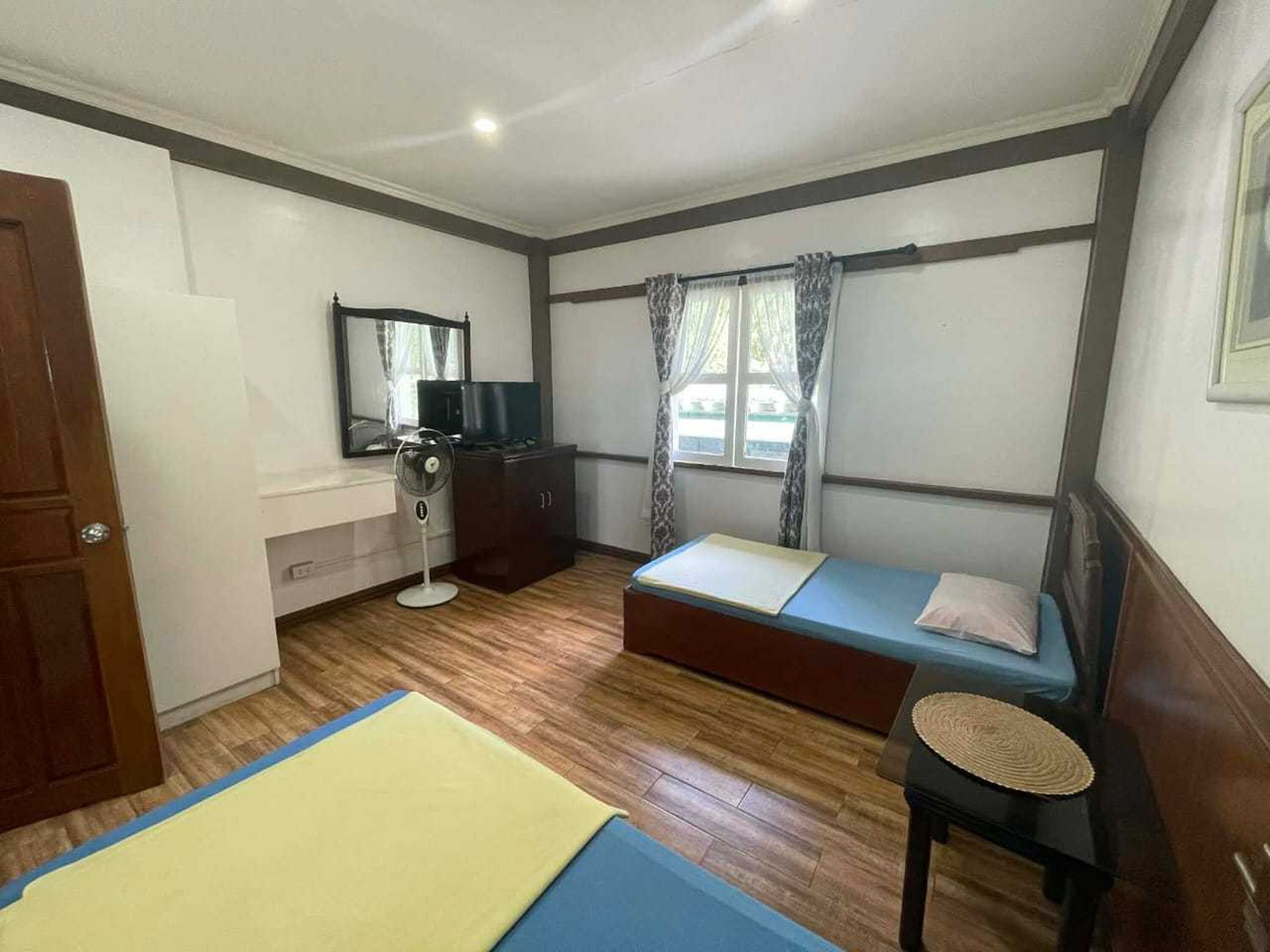 A small, well-lit room with two single beds, a desk with a chair, a television, and a window with curtains.