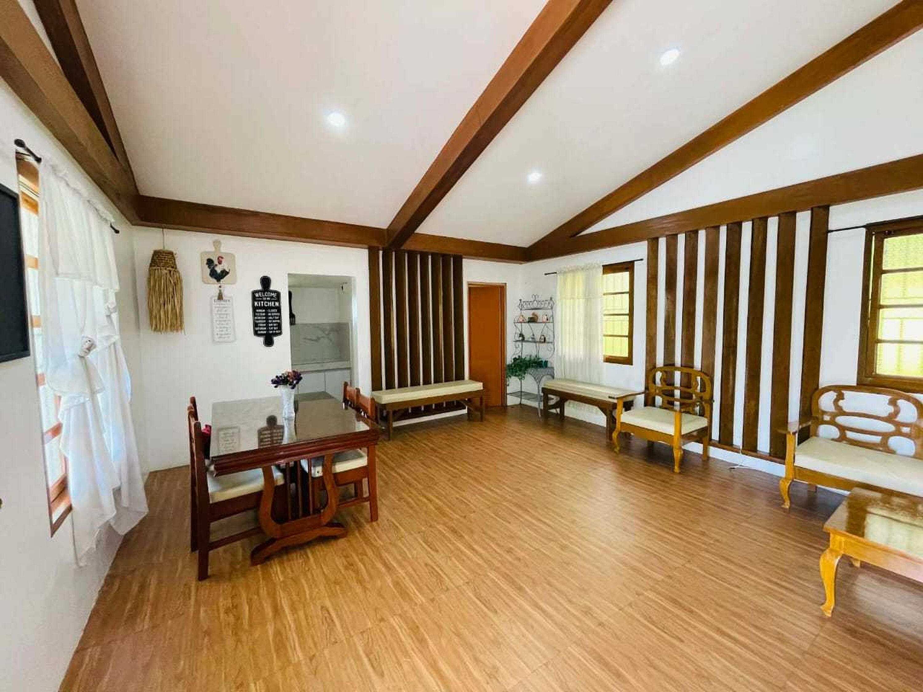 A spacious room with wooden flooring, a dining table with chairs, a sofa, and a bench. There are two windows with curtains, a wall-mounted sign, and a door.