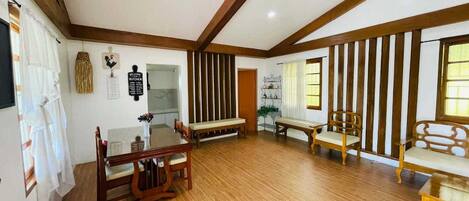 A spacious room with wooden flooring, a dining table with chairs, a sofa, and a bench. There are two windows with curtains, a wall-mounted sign, and a door.