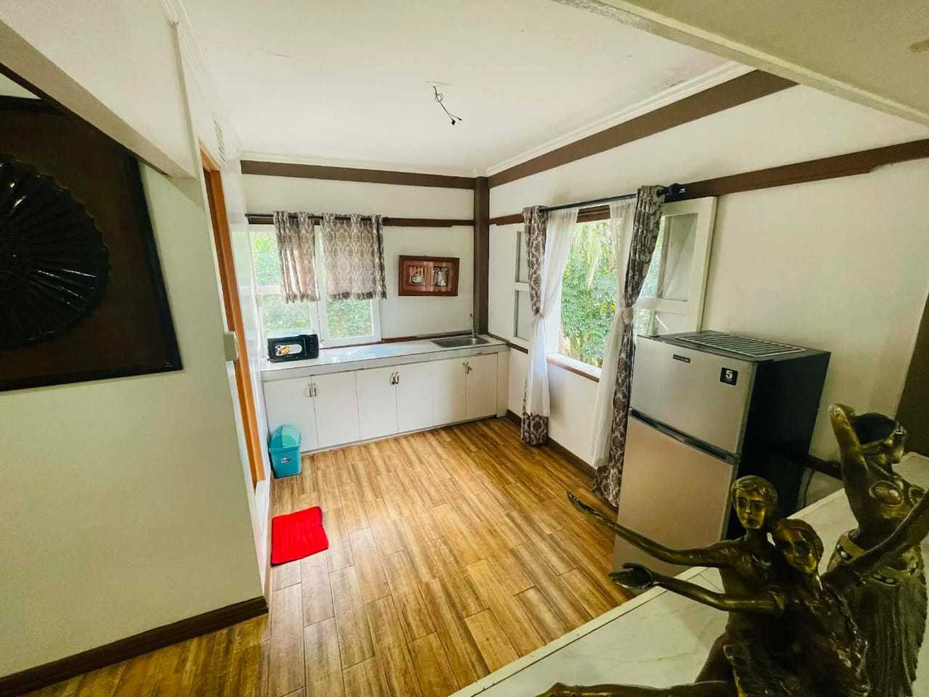 A compact kitchen with wooden flooring, a refrigerator, a sink, and a window with curtains.