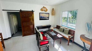 A room with a wooden door, a sofa with cushions, a table with a vase, a fan, and a window with a view of greenery. - 1193 Pinehall Farm Rizal (Antipolo)