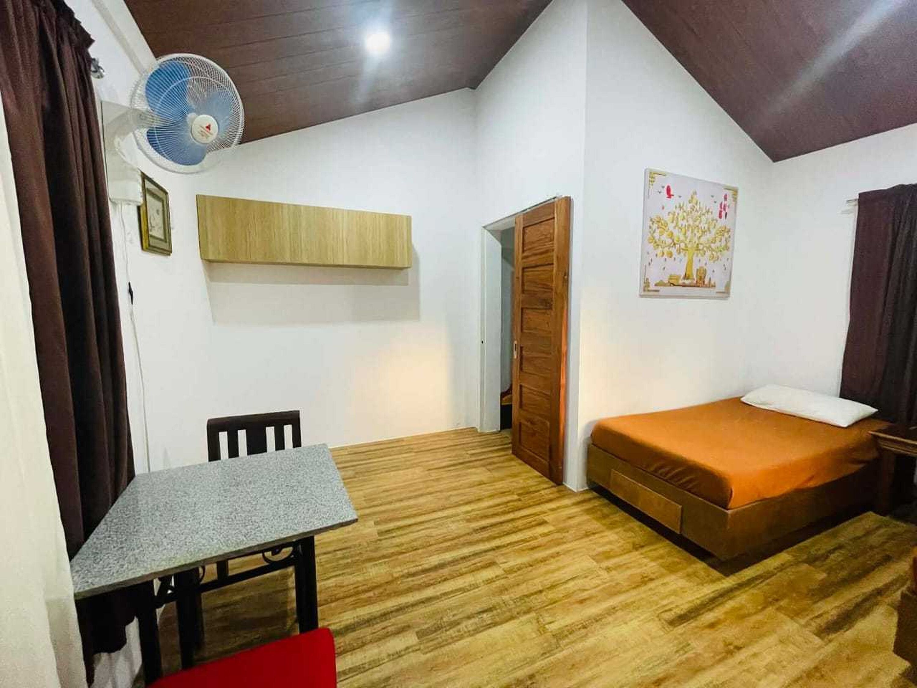 Quarto pequeno e bem iluminado, com piso de madeira, uma cama de solteiro, uma mesa com cadeira, um ventilador e uma porta que dá para outro cômodo.