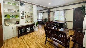 A spacious room with a wooden floor, a dining table with chairs, a white cabinet with decorative items, and a large window with curtains. - 1193 Pinehall Farm Rizal (Antipolo)