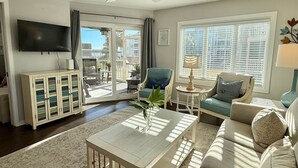 A spacious living room with a ceiling fan, a flat-screen TV, a coffee table with a vase, and a large sofa. - Newly Renovated First-Floor End-Unit Condo/Ocean Views/Beach Access/Free Parking (Isle of Palms)