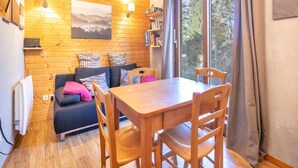 A cozy room with a wooden floor, a black sofa with plaid cushions, a wooden dining table with chairs, a bookshelf, and a large window with curtains. - Apt. in Chamrousse near supermarket (Chamrousse)