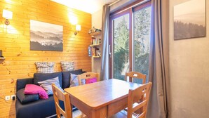 A cozy room with a wooden wall, a sofa with cushions, a table, chairs, a bookshelf, and a window with a view of trees. - Apt. in Chamrousse near supermarket (Chamrousse)
