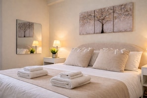 A neatly made bed with folded towels, a bedside lamp, a small plant, and a wall-mounted painting of a tree.