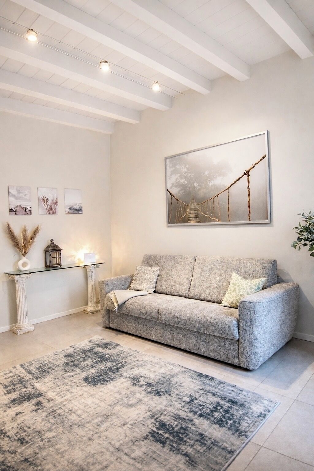 Una sala de estar con un sofá gris, un tapete estampado, una mesa de centro de vidrio y un cuadro de un puente.