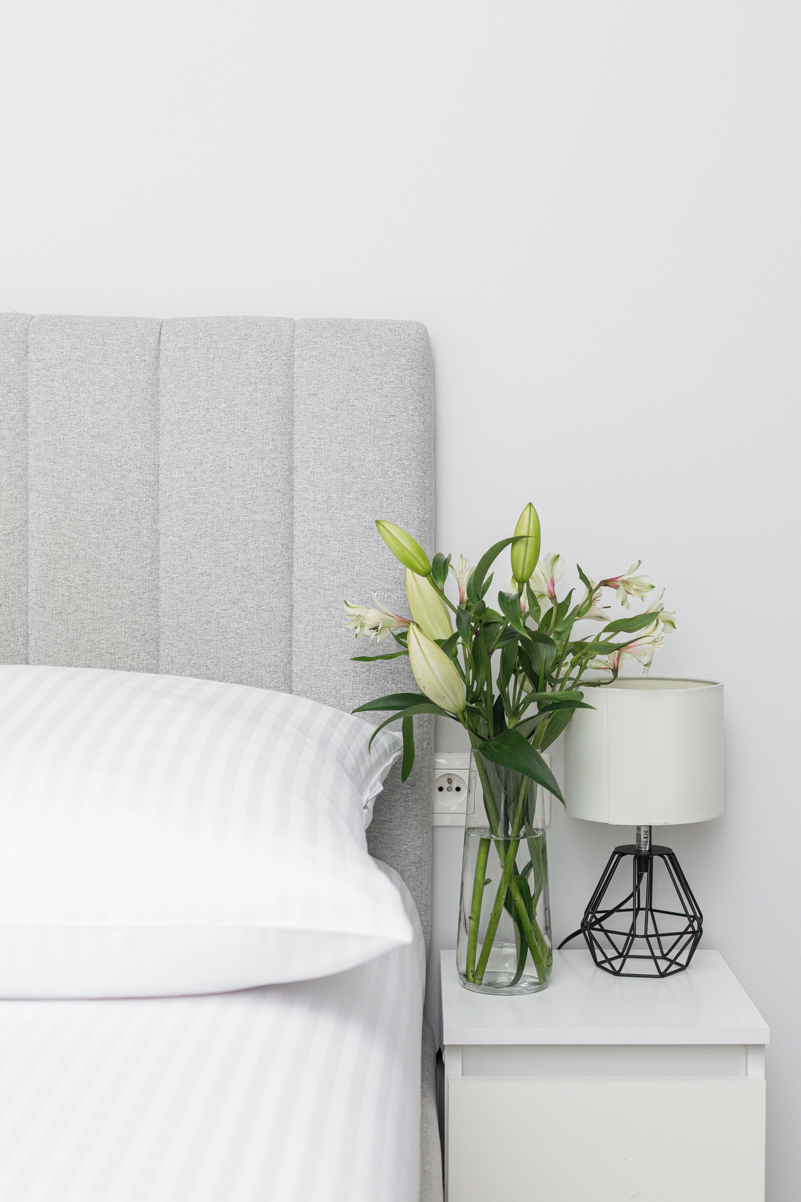A neatly made bed with a striped white duvet, a grey headboard, a bedside table with a clear vase of green and white flowers, and a black geometric lamp.