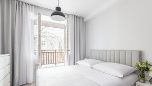 Een moderne slaapkamer met een groot bed, witte beddengoed, een grijs hoofdeinde, een nachtkastje met een vaas bloemen en uitzicht op een balkon met een balustrade.