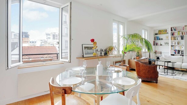 Una sala de estar moderna con una mesa de comedor de vidrio, sofás blancos, una estantería y un ventanal con vista a la ciudad.