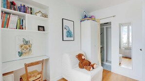 Una habitación con una silla blanca, un sofá blanco con un peluche de oso, una estantería llena de libros, un cuadro enmarcado de un loro y una puerta que da a otra habitación.