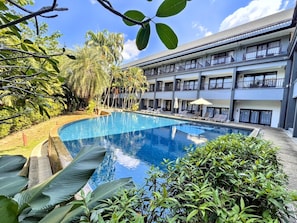 Un hotel con piscina, rodeado de exuberante vegetación y un edificio de varios pisos con balcones.