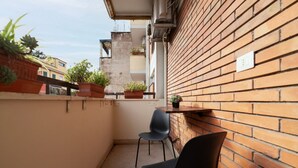 Un balcón estrecho con plantas en macetas, dos sillas negras y un muro de ladrillo.