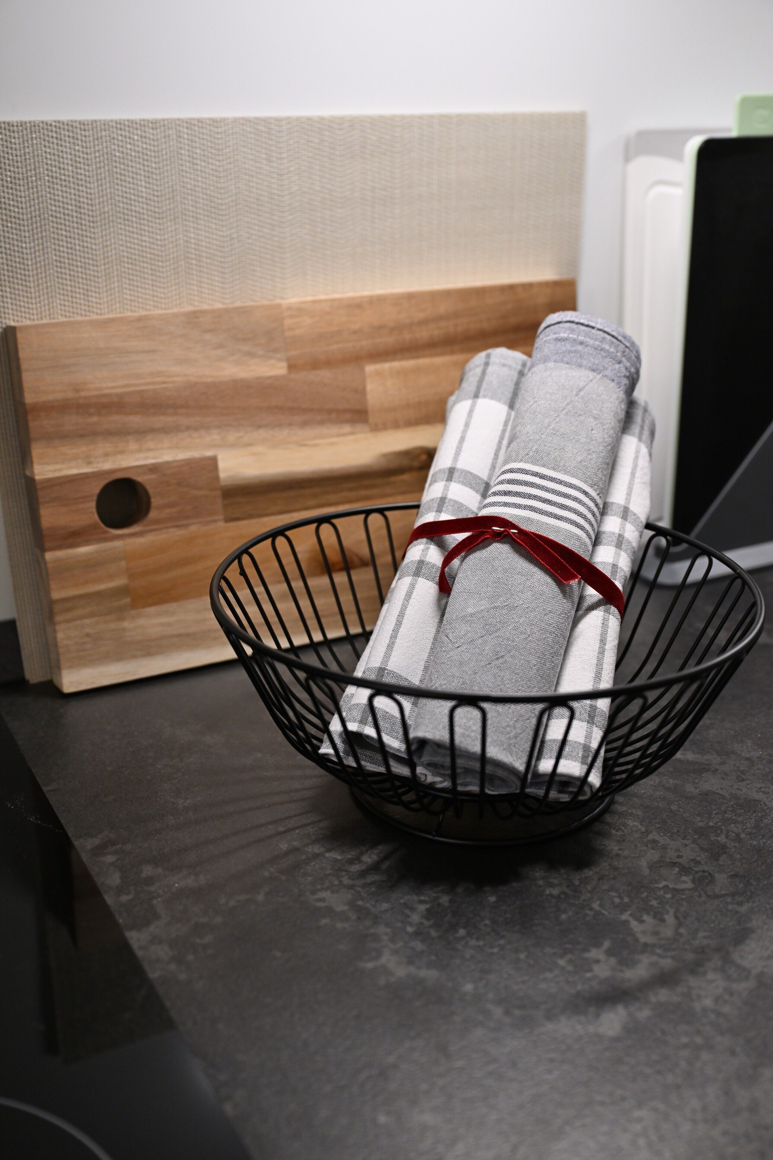 A kitchen counter with a cutting board, a metal basket with a rolled-up towel, and a white appliance in the background.