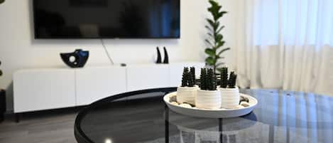 A modern living room with a glass coffee table, a flat-screen TV mounted on the wall, a white cabinet, and a potted plant.