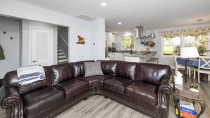 A living room with a dark brown leather sectional sofa, a staircase, a kitchen with white cabinets, and a dining area with a table and chairs. - Modern Huntersville Oasis w Sauna, Fire Pit, Screened Patio & Fenced Yard For Pets! + 15% Off Stay! (Huntersville)