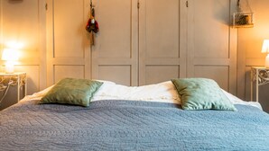 Un lit avec une couette bleue, deux oreillers, une table de chevet avec une lampe et une armoire dans laquelle est accroché un manteau.