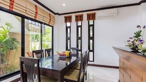 A dining area with a glass table, wooden chairs, and a fruit bowl. There are three windows with wooden frames and an air conditioning unit on the wall. - Baan Narakorn (Krabi)