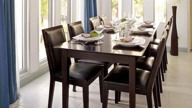 A dining area with a long table set for six, surrounded by dark wooden chairs. Large windows with blue curtains allow natural light to fill the space. - Baan Gecko (Krabi)