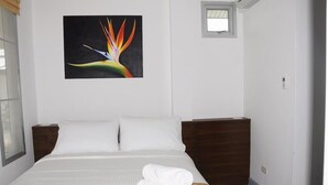 A hotel room with a bed, white linens, a wooden nightstand, a small window, and a wall-mounted painting of a bird of paradise. - Baan Gecko (Krabi)