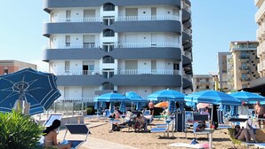 Um hotel com vários andares, varandas, área de piscina com espreguiçadeiras e guarda-sóis, e uma praia de areia.