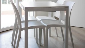 Une table à manger blanche, accompagnée de quatre chaises, se trouve dans une pièce dotée d’une porte-fenêtre coulissante qui donne sur un balcon.