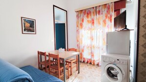 Een kleine, schone kamer met een bed, een tafel met stoelen, een wasmachine en een raam met gordijnen.