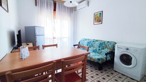 A small, bright room with a wooden dining table, chairs, a sofa, a washing machine, and a television. - Cozy flat near the beach (Lido di Jesolo)